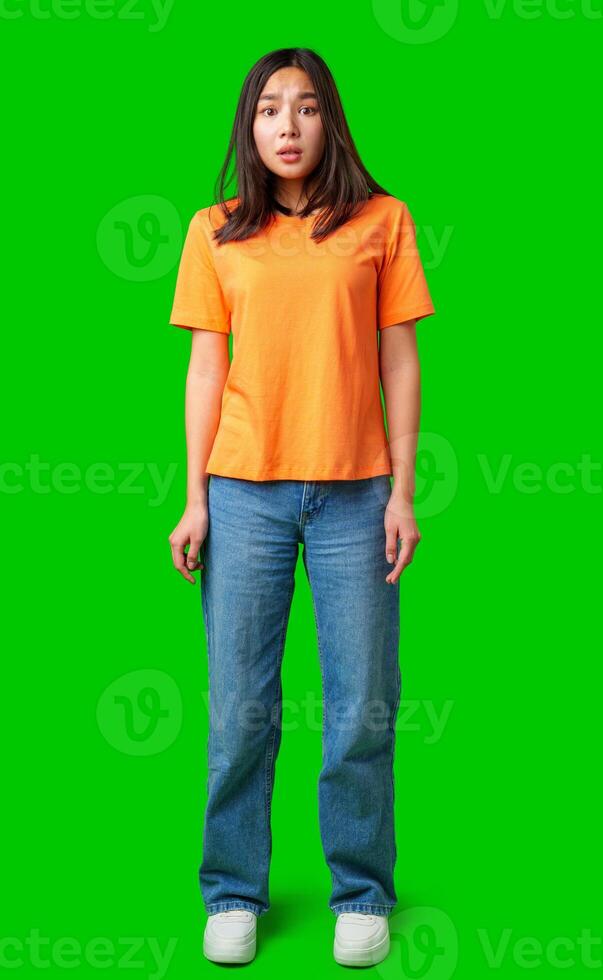 Young woman standing on green background wearing an orange shirt and blue jeans looking surprised photo