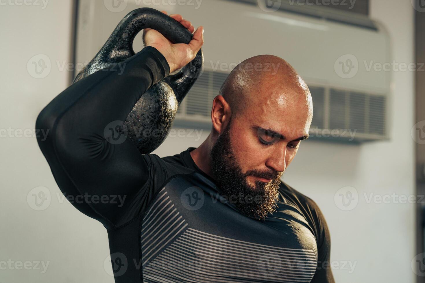 Bald man lifting kettlebell with focused expression in a gym setting during workout session photo