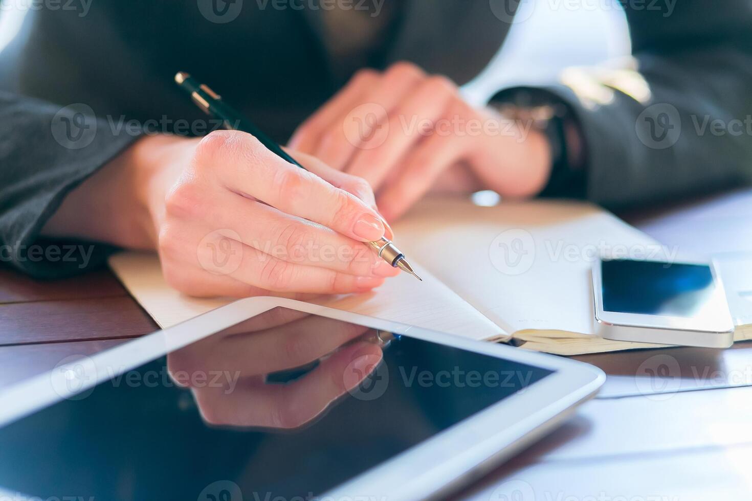 Person writing notes with a pen while seated at a desk with a tablet and smartphone nearby in a well-lit space photo