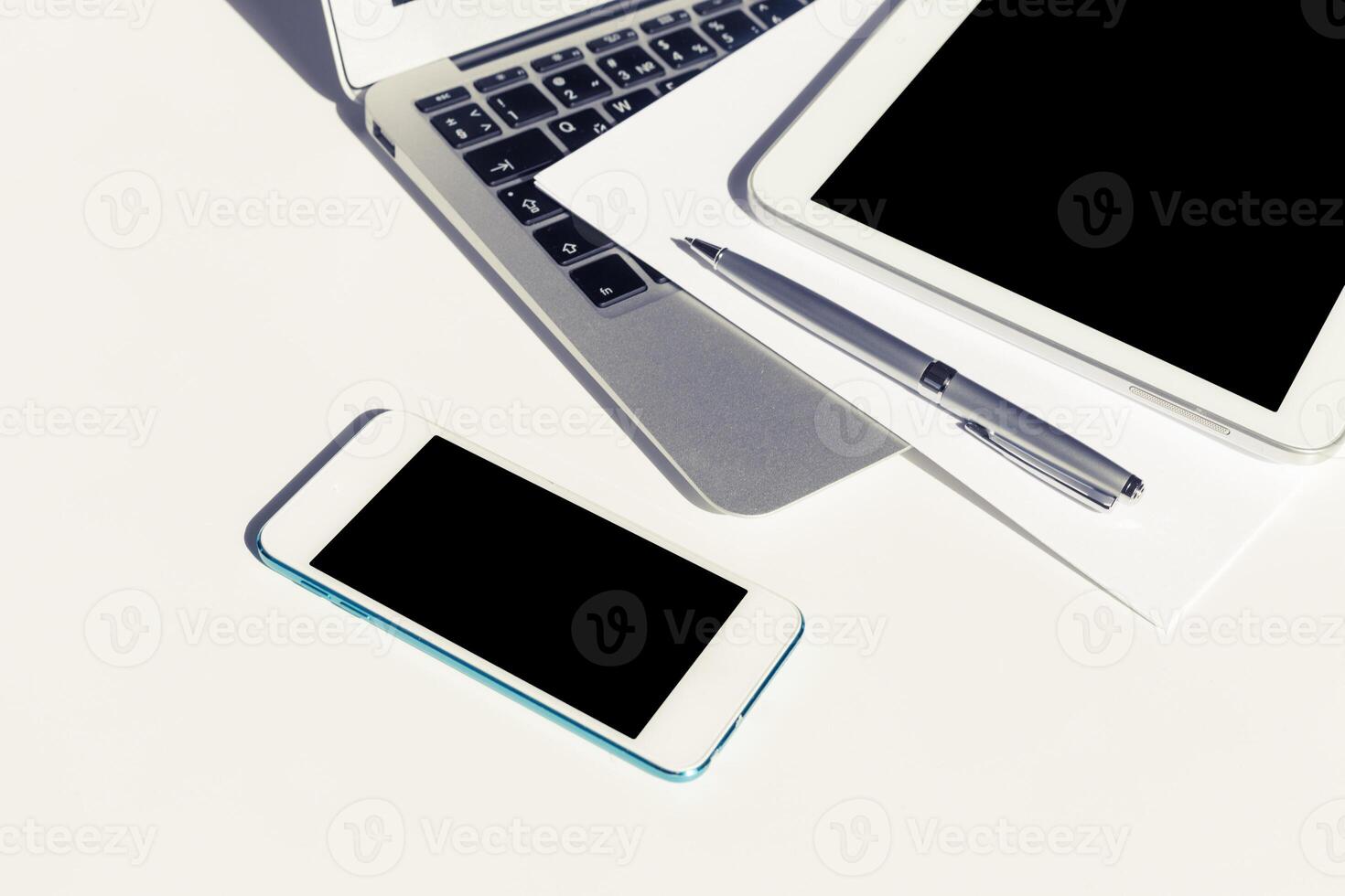 Workspace with electronic devices including smartphone, tablet, and laptop on a white surface during daylight hours photo