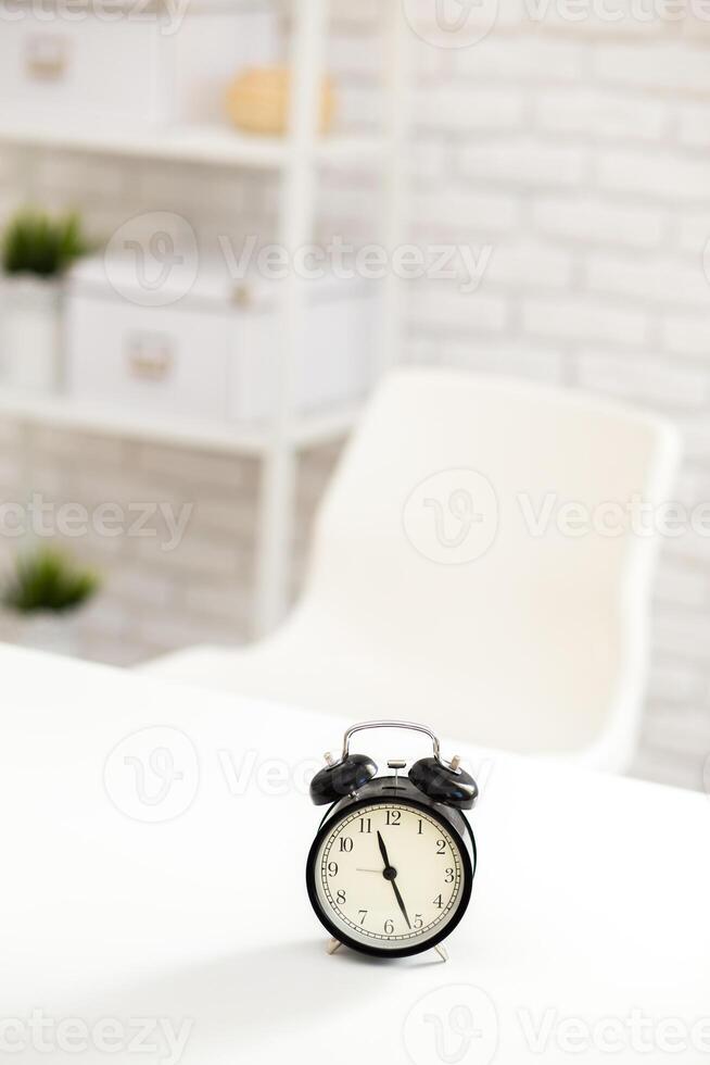 Clock placed on a table in a minimalistic workspace with a white chair and organized shelves photo