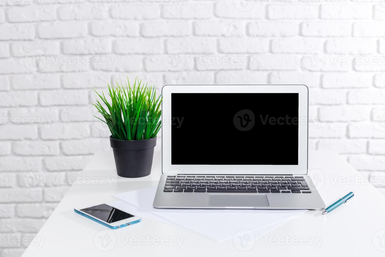 Modern workspace setup with laptop, smartphone, and decorative plant on a white table against a brick wall photo