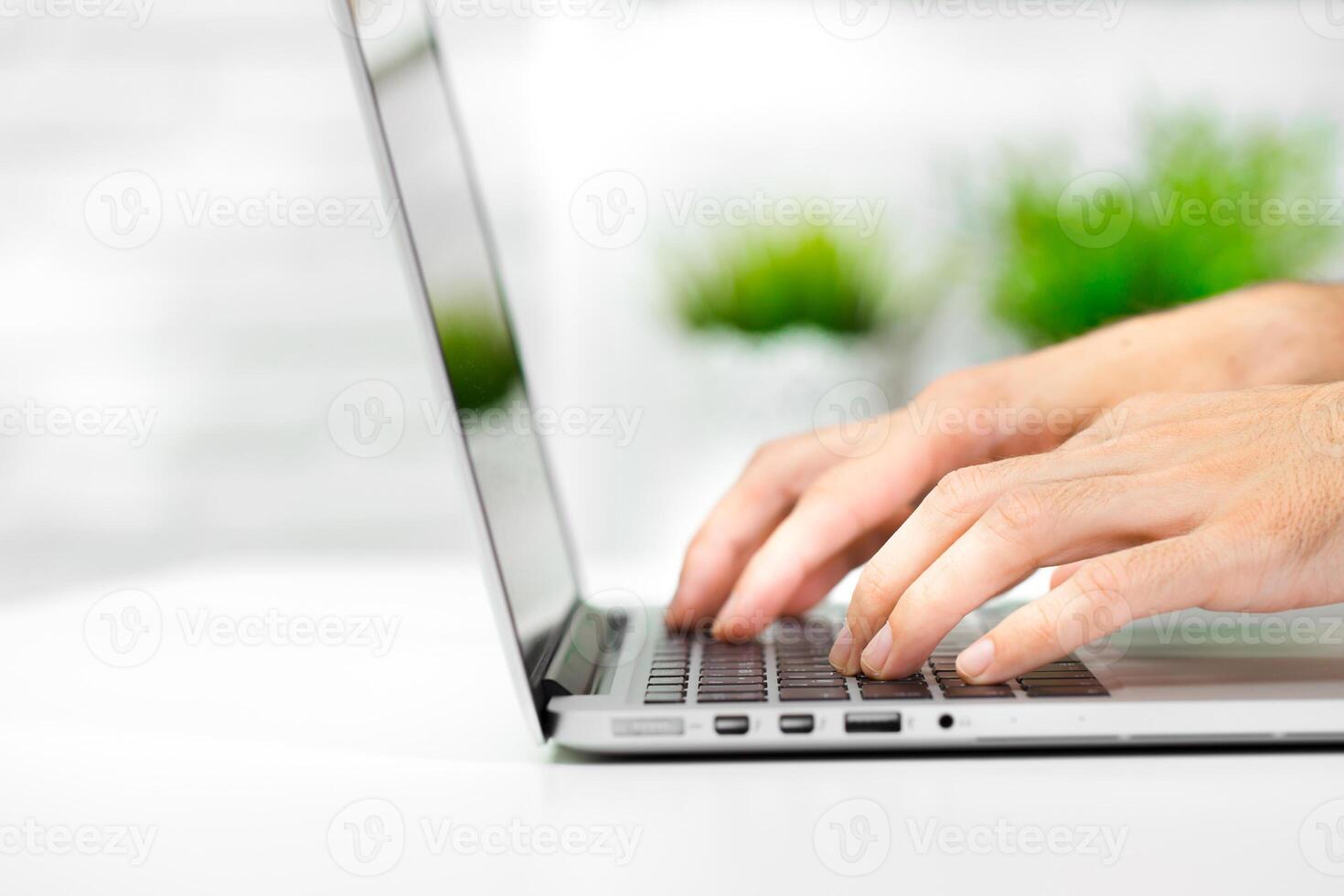 Hands typing on a laptop in a well-lit workspace with green plants in the background during daytime photo