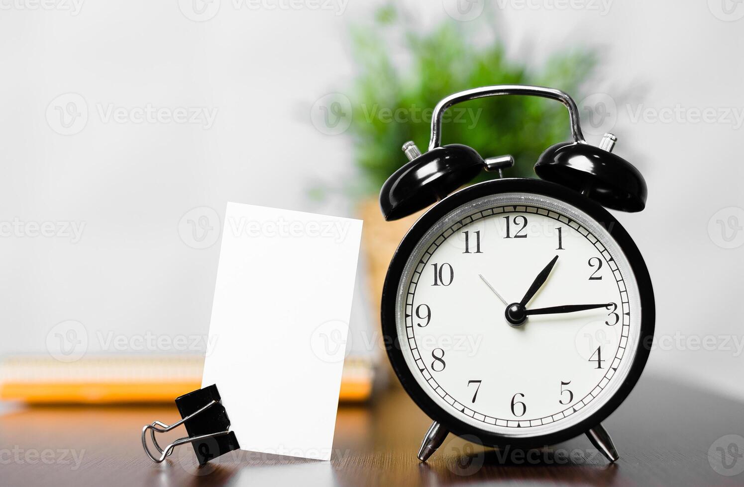 Classic black alarm clock on wooden desk with blank card and binder clip, showcasing workspace organization photo