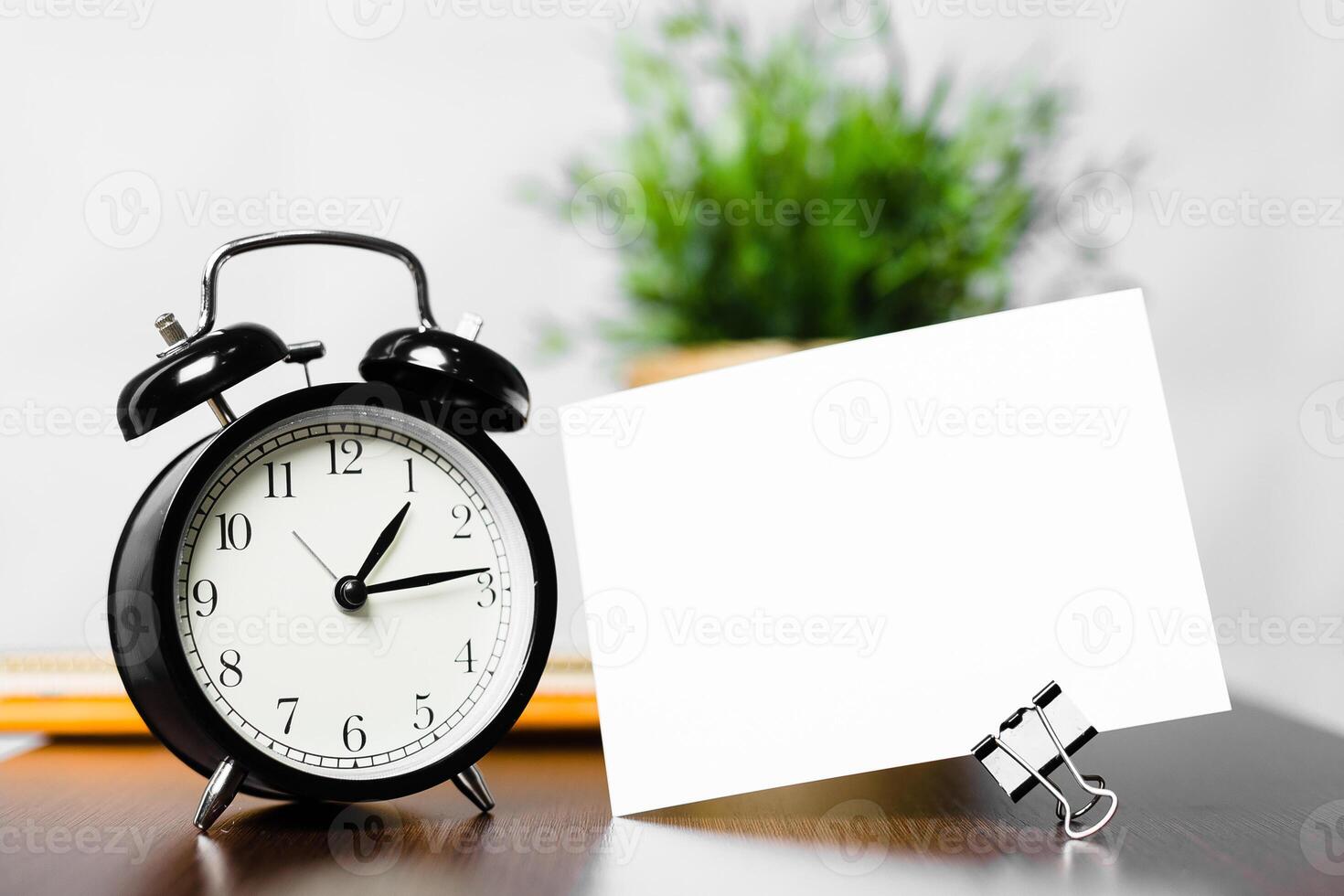 Black alarm clock and blank card on desk beside green plant in a modern workspace photo