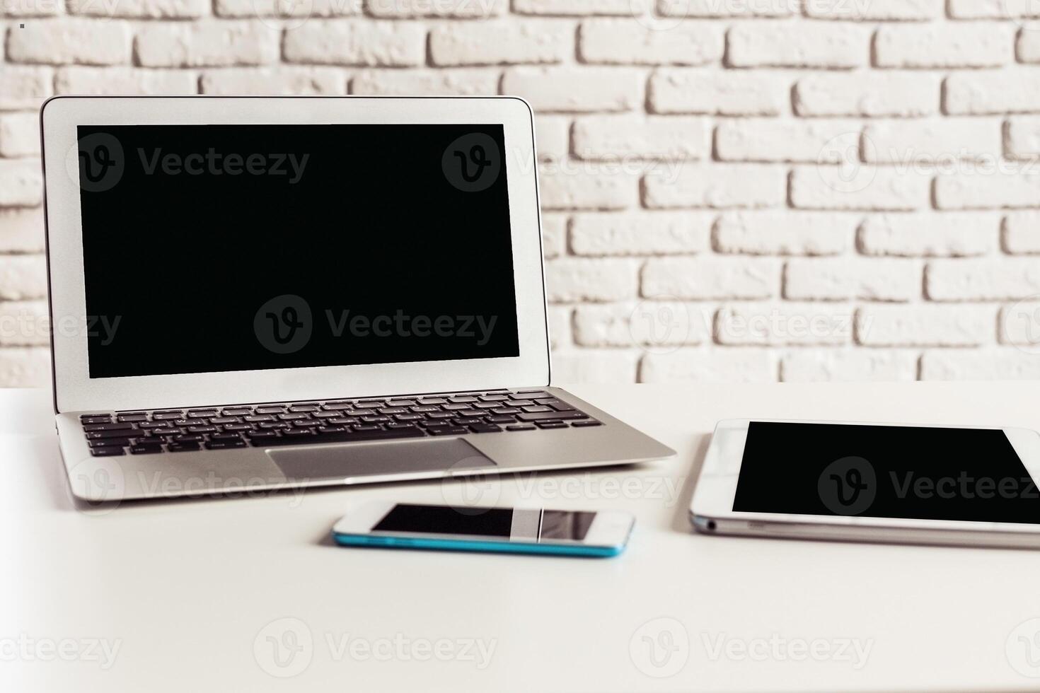 Workspace featuring a laptop, smartphone, and tablet on a white brick background during the afternoon photo