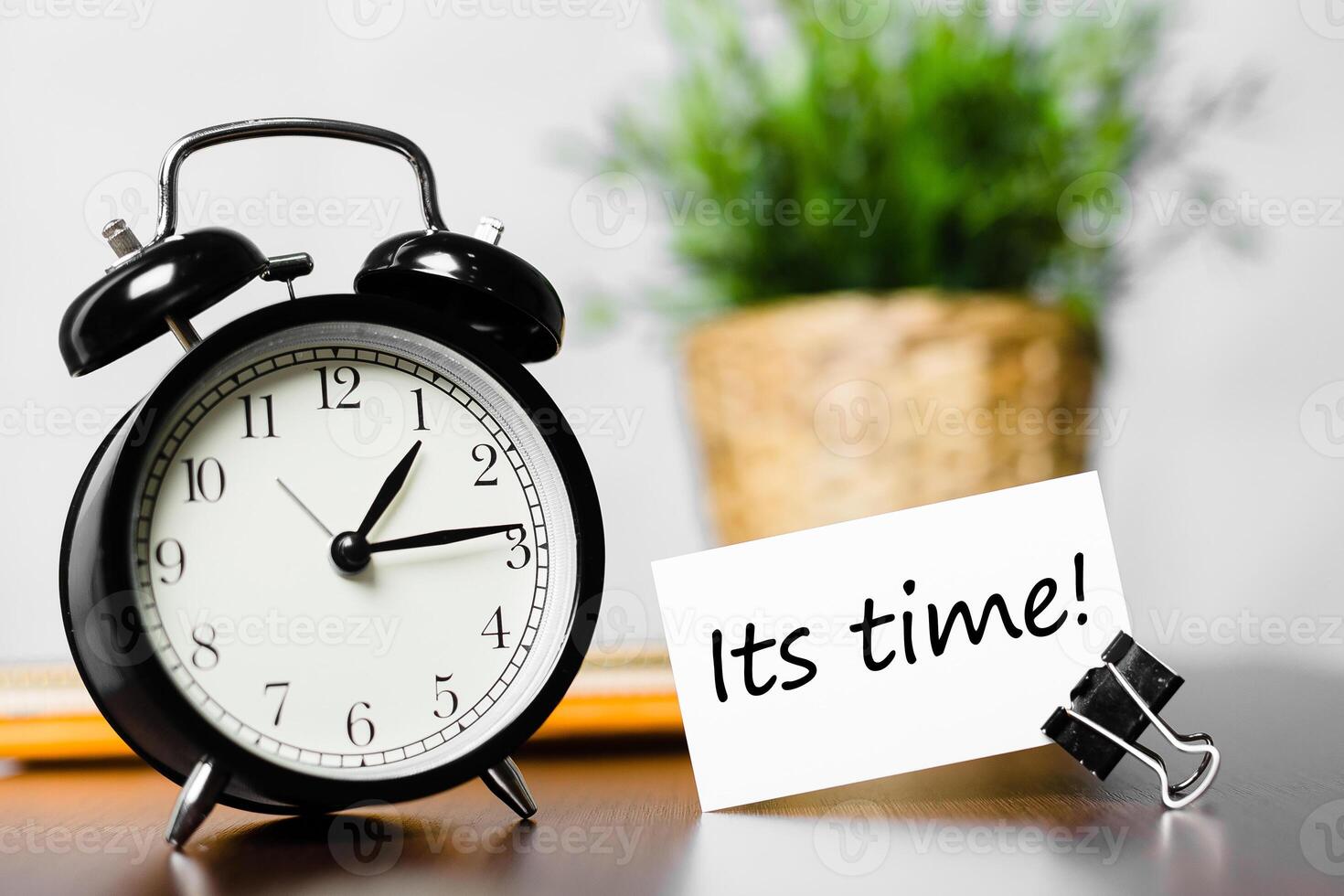 Black alarm clock next to a sign reading its time on a wooden background with a plant photo