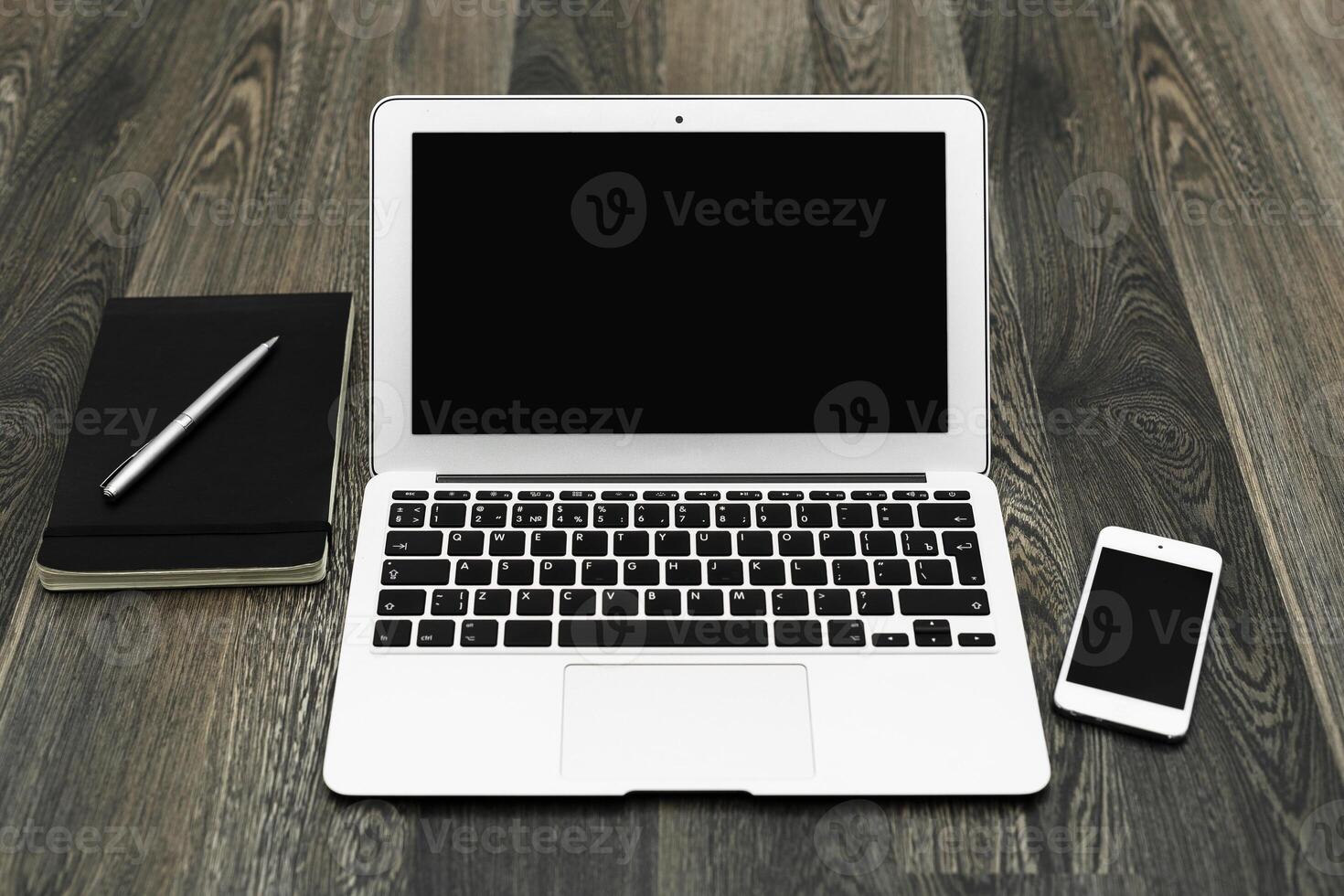 Workspace setup featuring a laptop, smartphone, notebook, and pen on a wooden desk during daylight hours photo