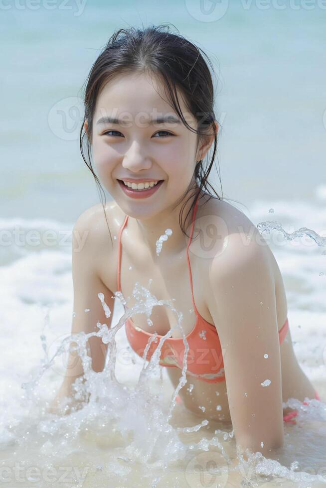 Young Asian girl, with light skin and long dark hair, smiling while sitting in shallow water. She is wearing a coral bikini, and water splashes around her photo
