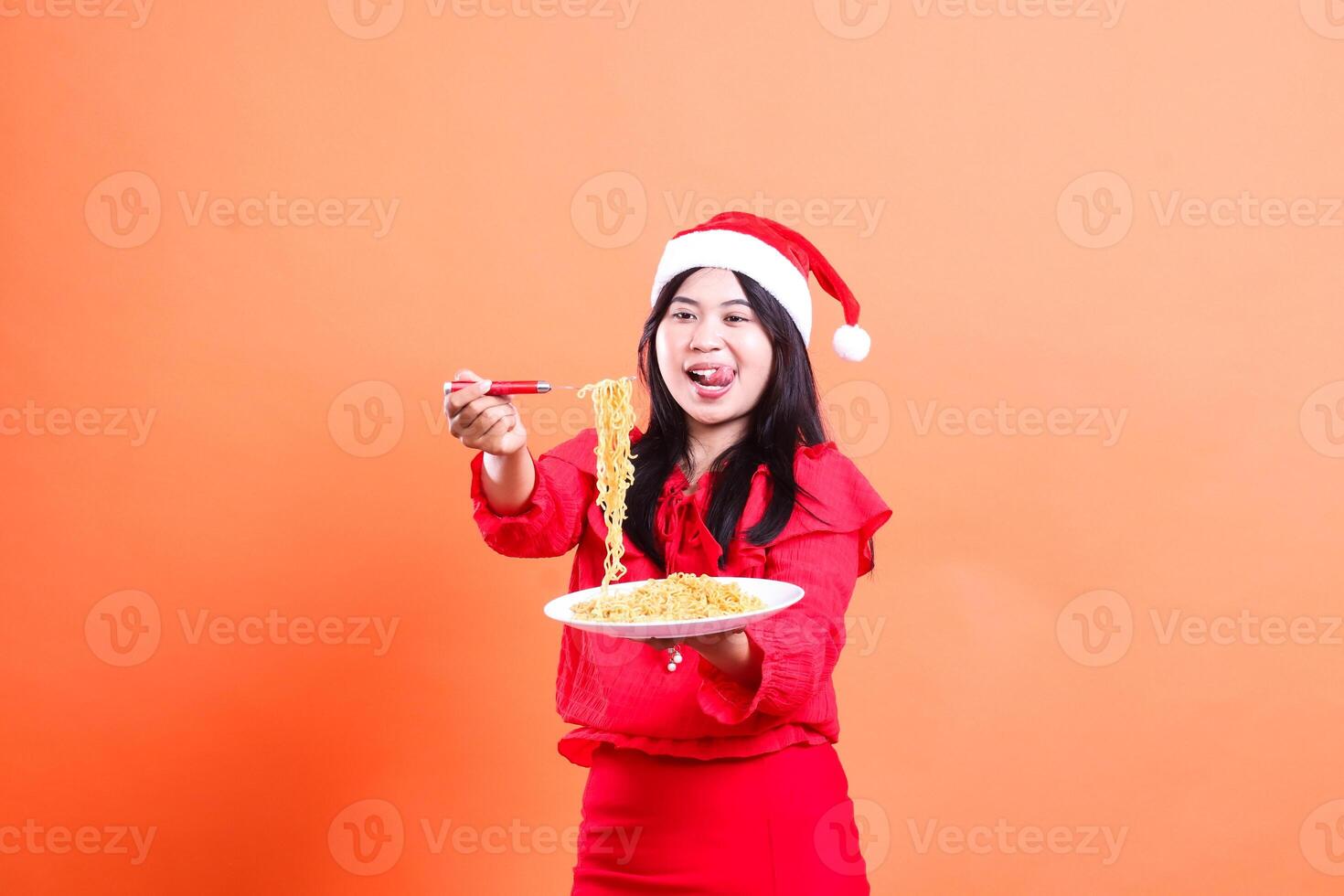 beautiful woman charm wearing christmas dress, santa hat, hungry looking at noodles, hand holding noodle fork and white plate filled with noodles in front, isolated on orange background photo