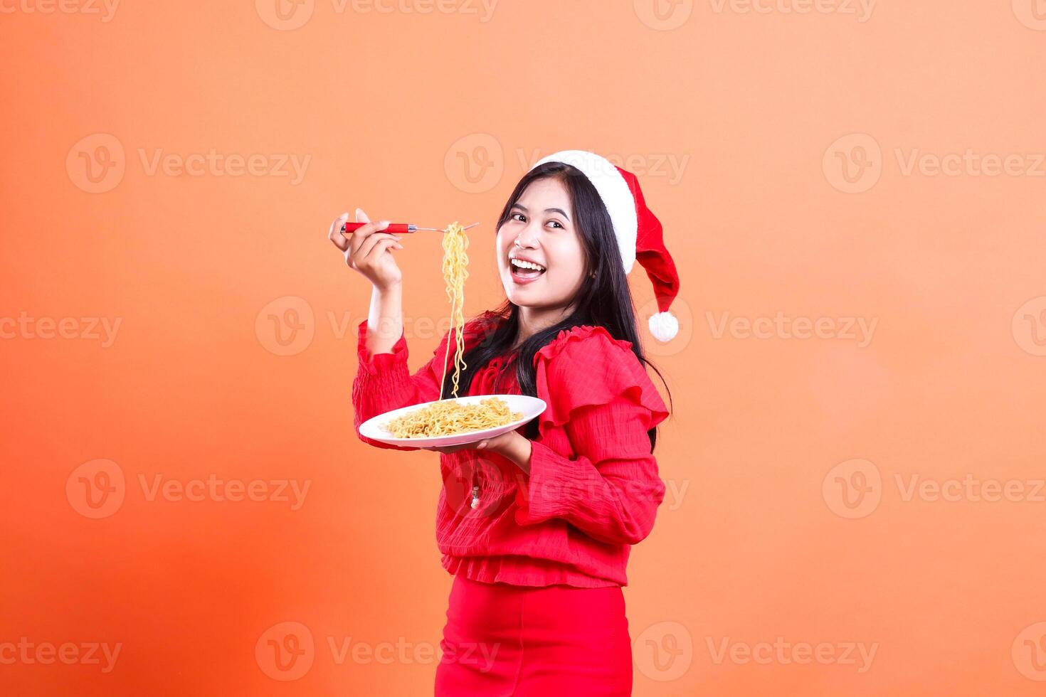 expression of ladies wearing christmas dress, santa hat, cheerful right sideways look left,hand holding fork filled with noodles upwards and white plate filled with noodles, isolated orange background photo