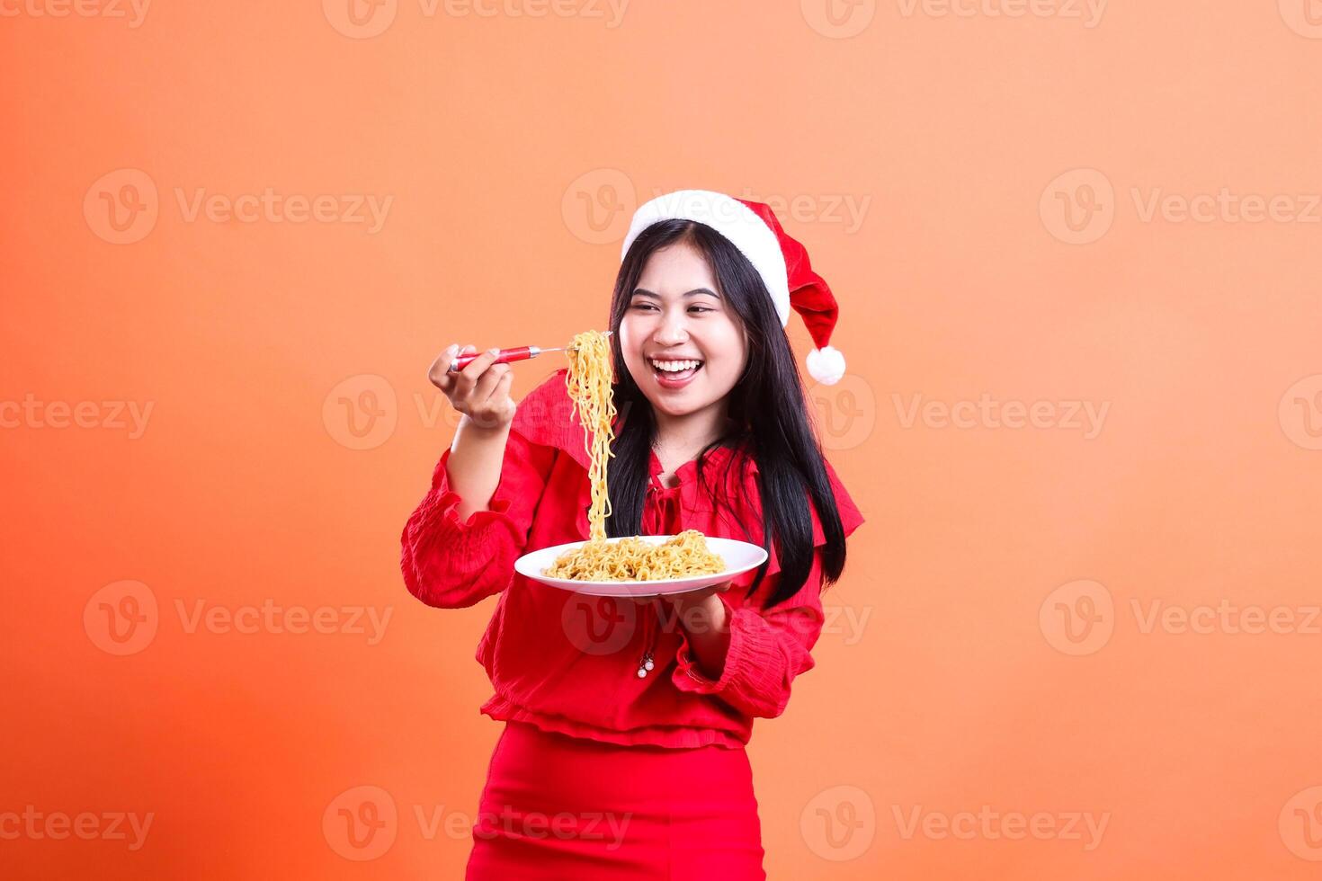 cheerful asian ladies looking at noodles, hand holding fork filled with noodles from white plate while looking at them, wearing red christmas blouse, Santa hat, isolated on orange background photo
