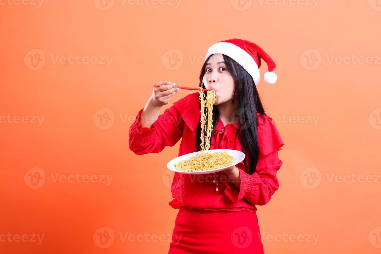 Asian adult woman surprised eating noodles in front, hand holding chopsticks to mouth and white plate filled with noodles, wearing red Christmas blouse, Santa hat, isolated on orange background photo