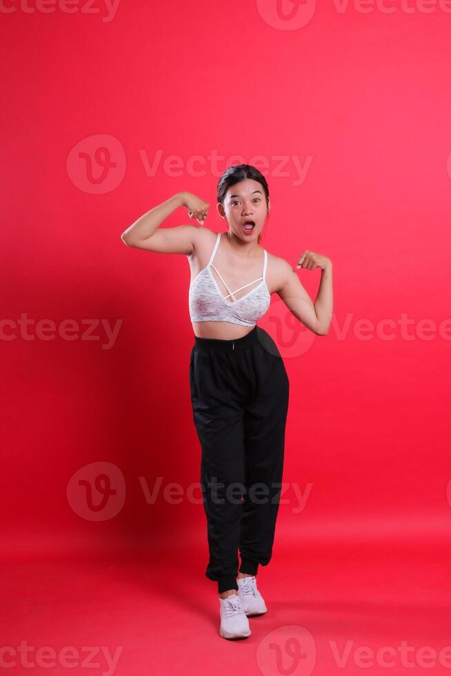 A woman in a sports bra and black pants is flexing her arms while standing with her mouth open. isolated on a red background photo