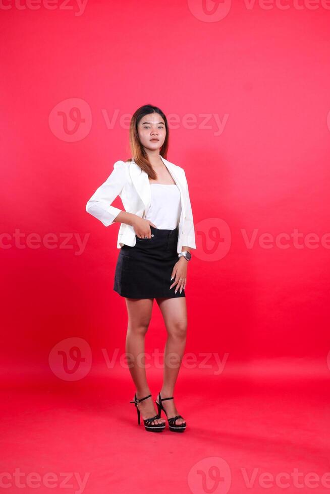 full body of beautiful young office female wearing elegant white suit with hands in skirt pocket and touching thighs, standing isolated on red background. for corporate, business, fashion content photo