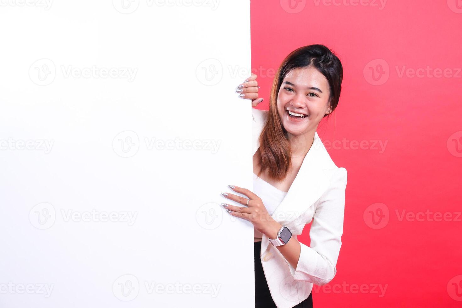 A smiling woman peeking behind a large blank white sign, she is wearing a white blazer and a smart watch and her nails are metallic, isolated on a red background photo