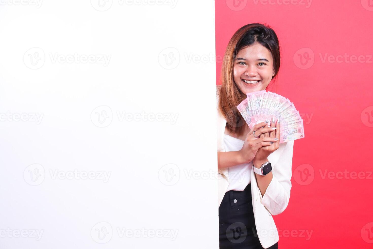 A woman holding a fan of banknotes, wearing a jacket and a watch behind a white panel, isolated on a red background photo