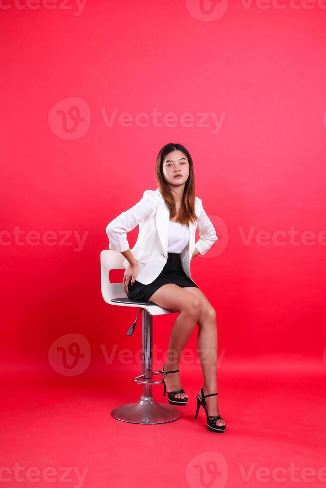 full body young female wearing elegant white suit seductively both hands hold waist, sitting on hydraulic chair isolated on red background. for corporate, business, fashion, lifestyle content photo