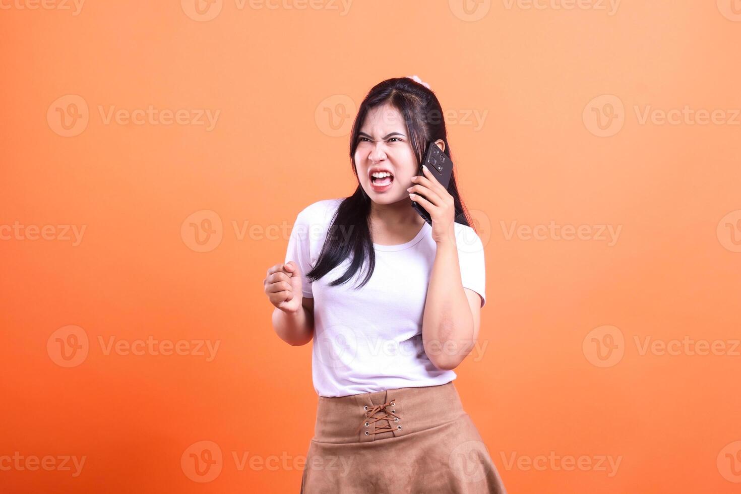 Angry woman on phone, with white t-shirt and brown skirt, isolated on orange background photo