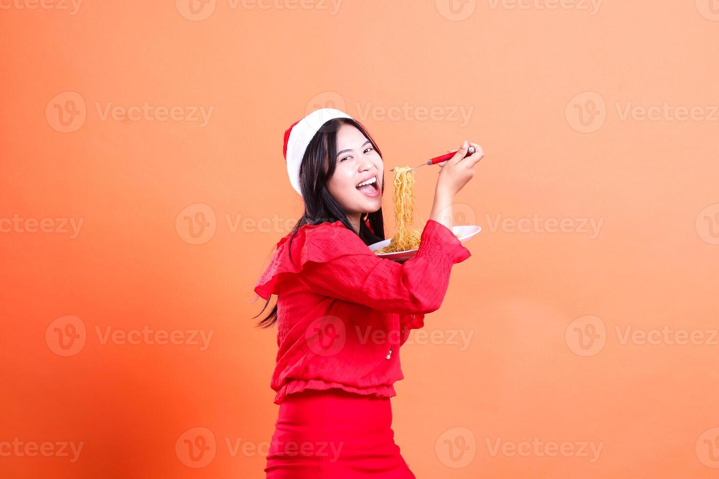 young indonesia woman slanted left laughing looking at camera, hand holding noodle fork and white plate filled with noodles, wearing red christmas blouse, Santa hat, isolated on orange background photo