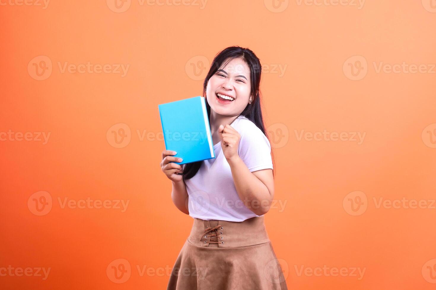 Young woman holding a blue book while smiling cheerfully with clenched fist isolated on orange background photo