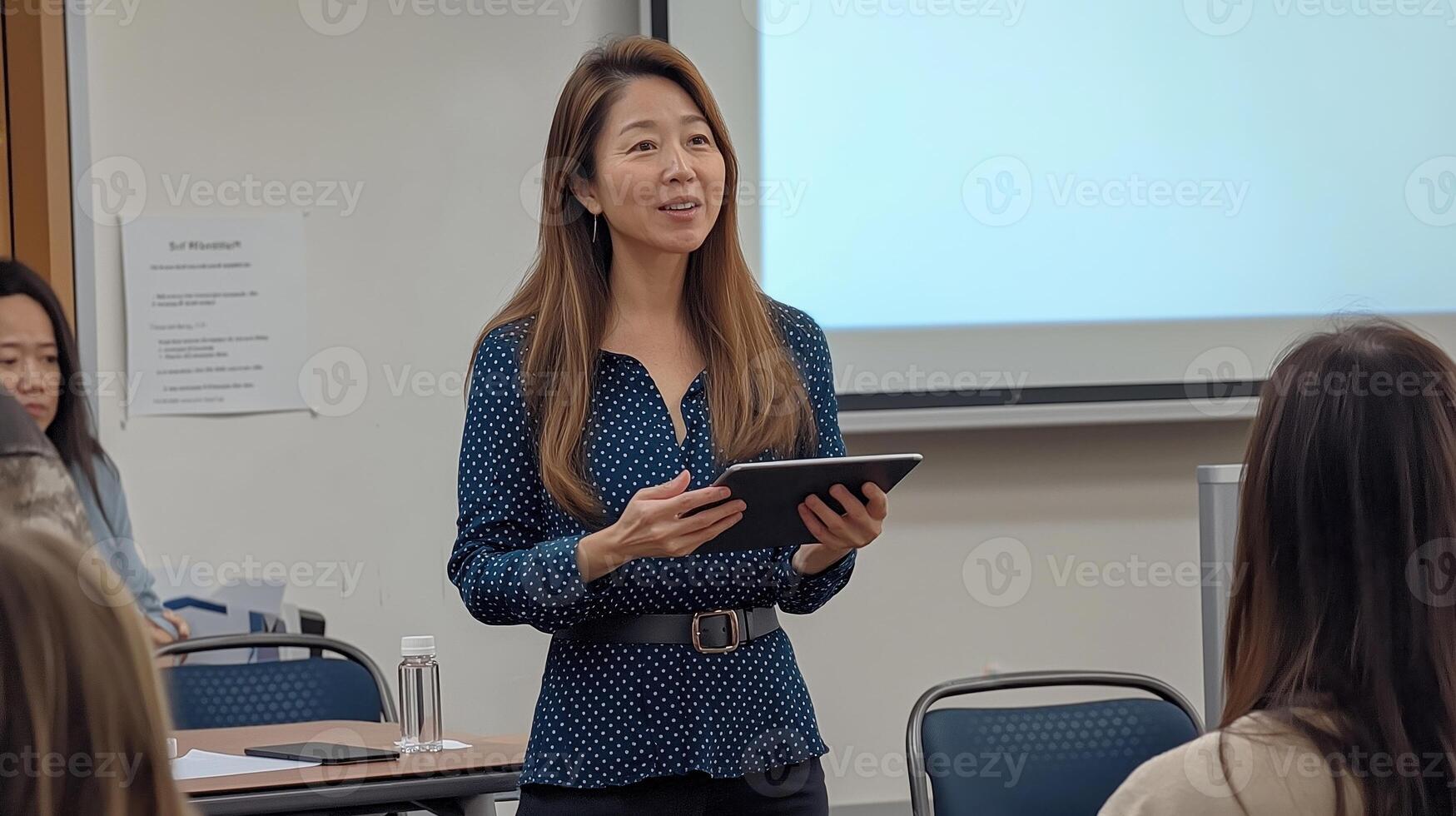 asian young woman standing in front of a group of people in a classroom. She is holding a tablet and appears photo