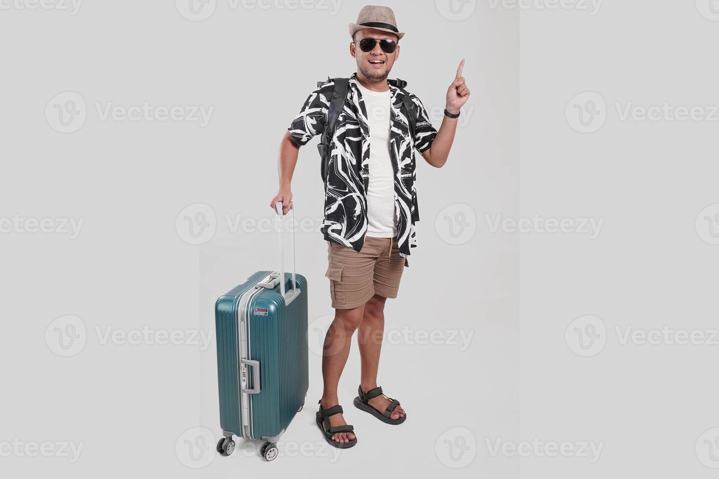 Happy young Asian man with suitcase wearing hat and glasses pointing to the copy space upward. photo