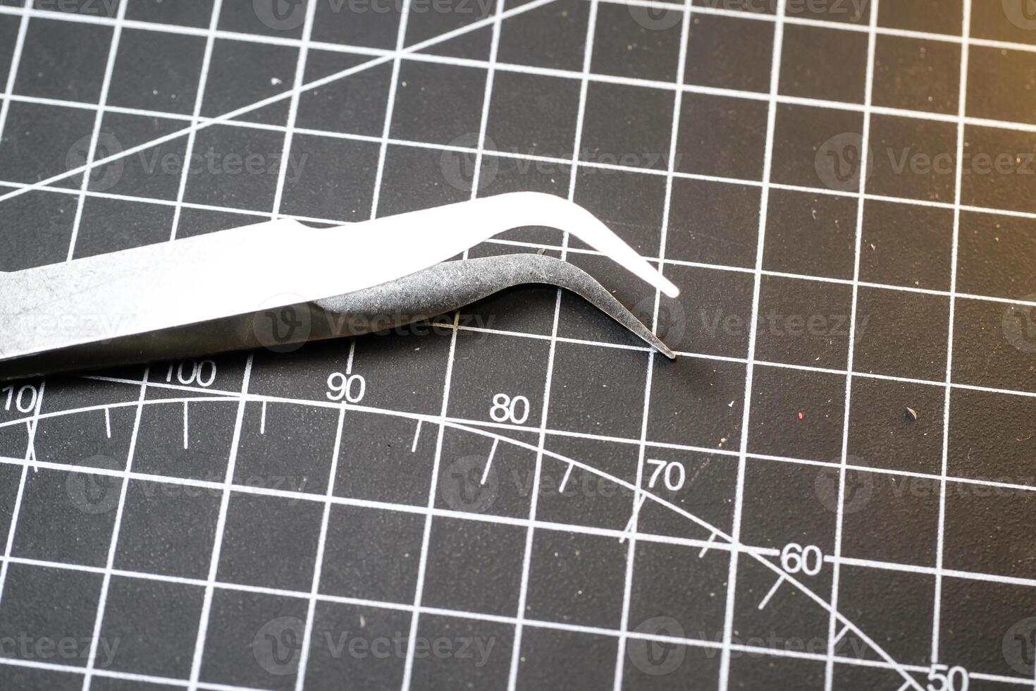 Metal precision tweezer with pointed tip. Tweezer is on a black cutting mat with lines. A tool that functions to clamp and pull out small objects. photo