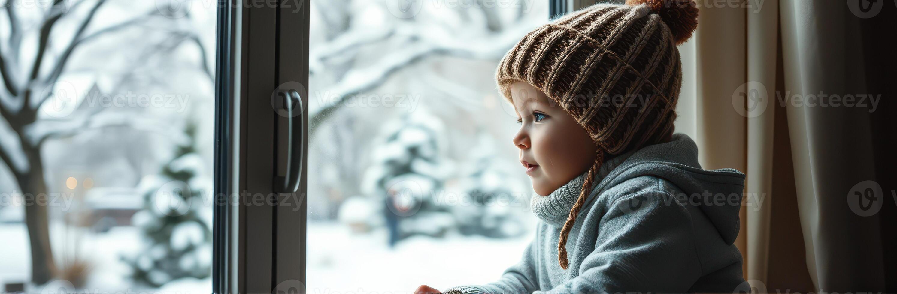 Child gazing out the window on a snowy winter day with a cozy hat and warm sweater photo