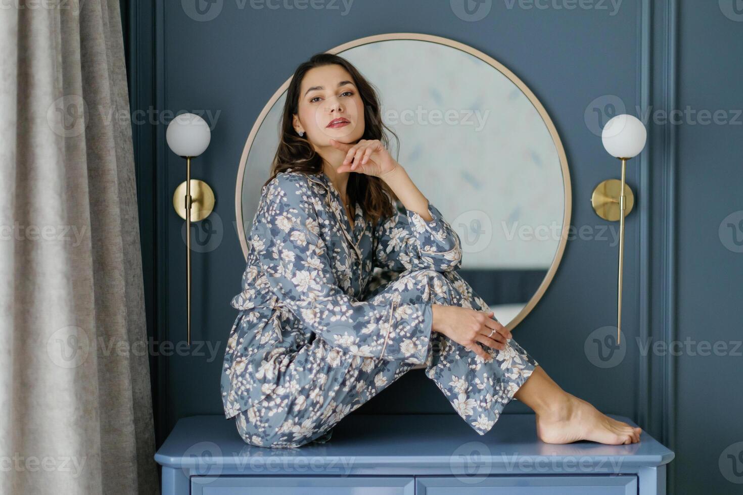 Woman wearing pajamas sitting on furniture in bedroom with round mirror and wall lamps. photo