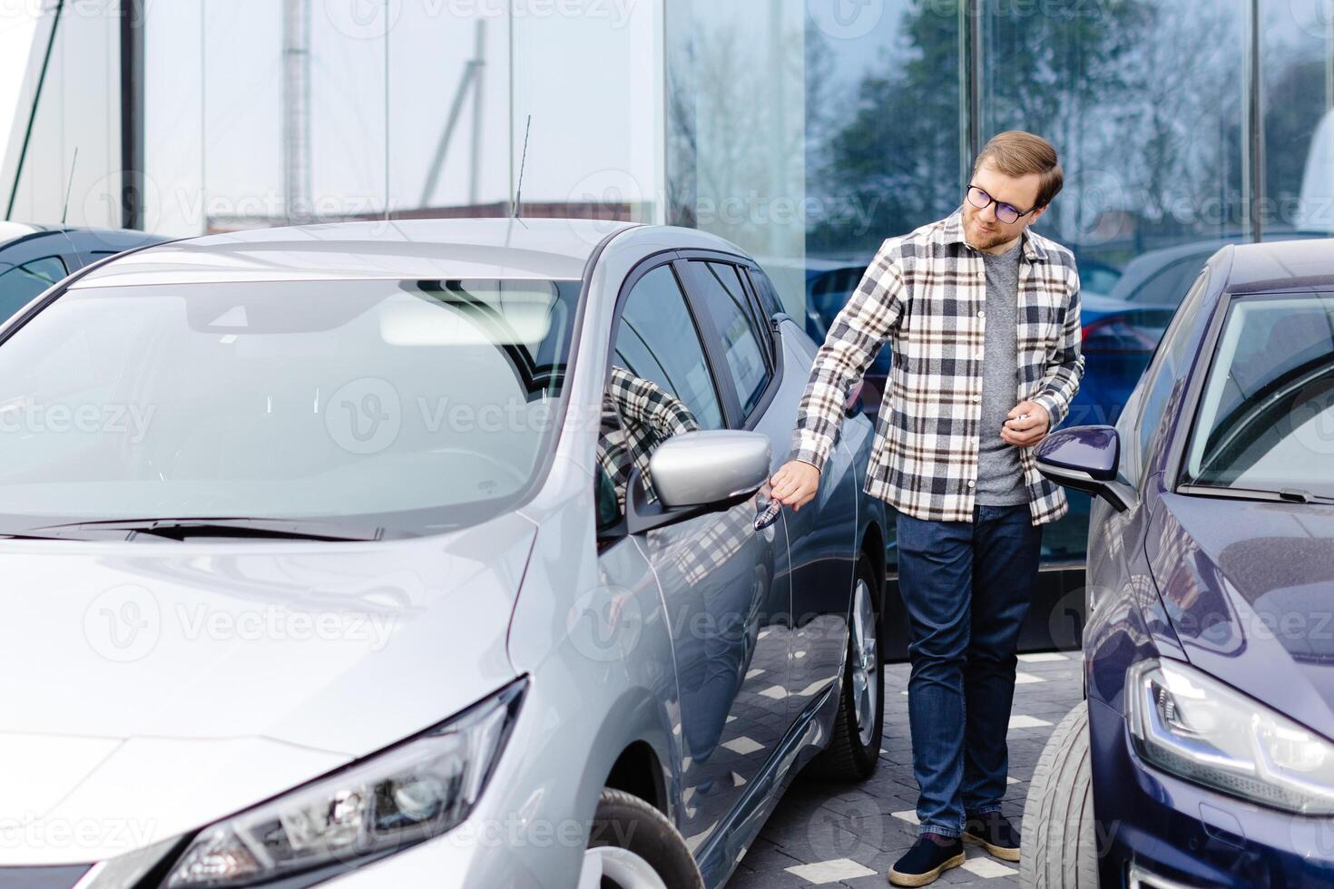Young bearded man embracing his new car at the dealership smiling joyfully. photo