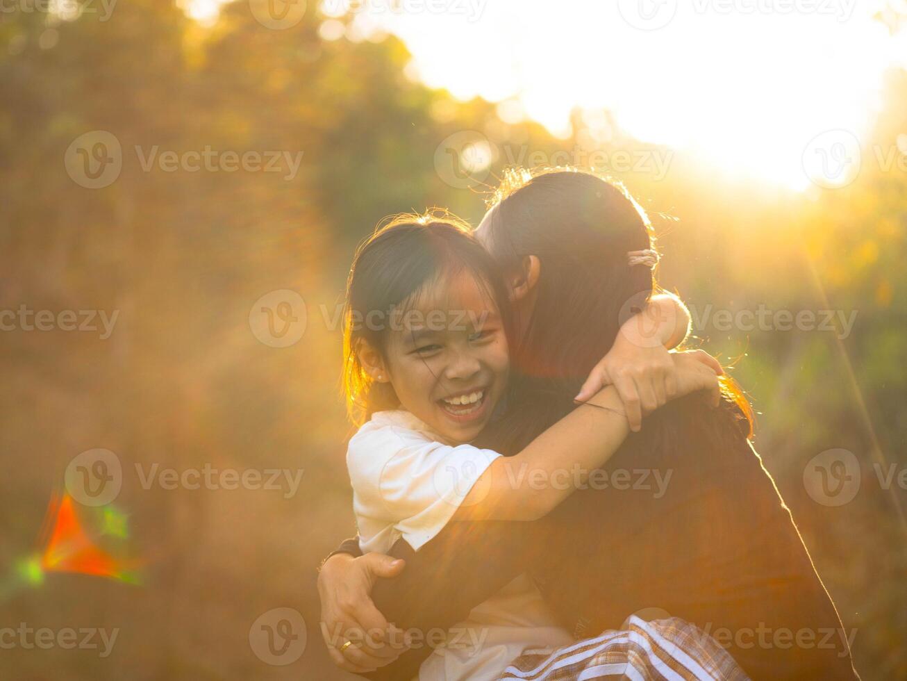 Mother and daughter are playing together in the park. Happy mother hugging her daughter sitting in the park with sunset light in nature. photo