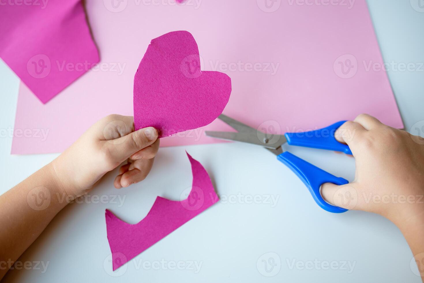 Child creating pink paper heart with blue scissors on craft project photo
