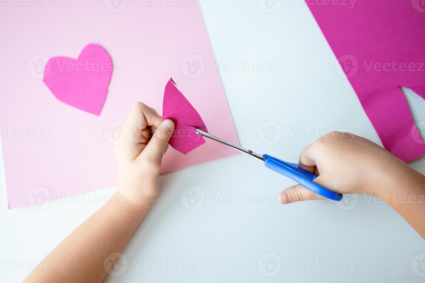 Child cutting pink paper heart with scissors on white table photo