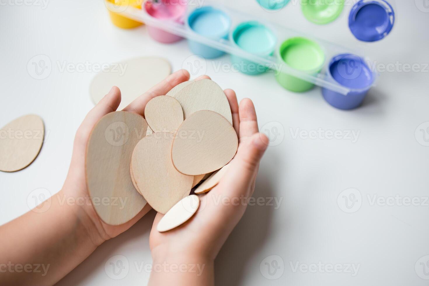 Child holding Easter wooden egg shapes with paints in the background photo