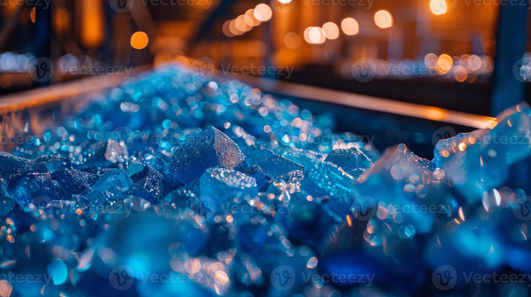 Sparkling blue glass fragments fill a conveyor belt in a factory illuminated by warm lights, showcasing the vibrant color and texture against the industrial backdrop at dusk. photo