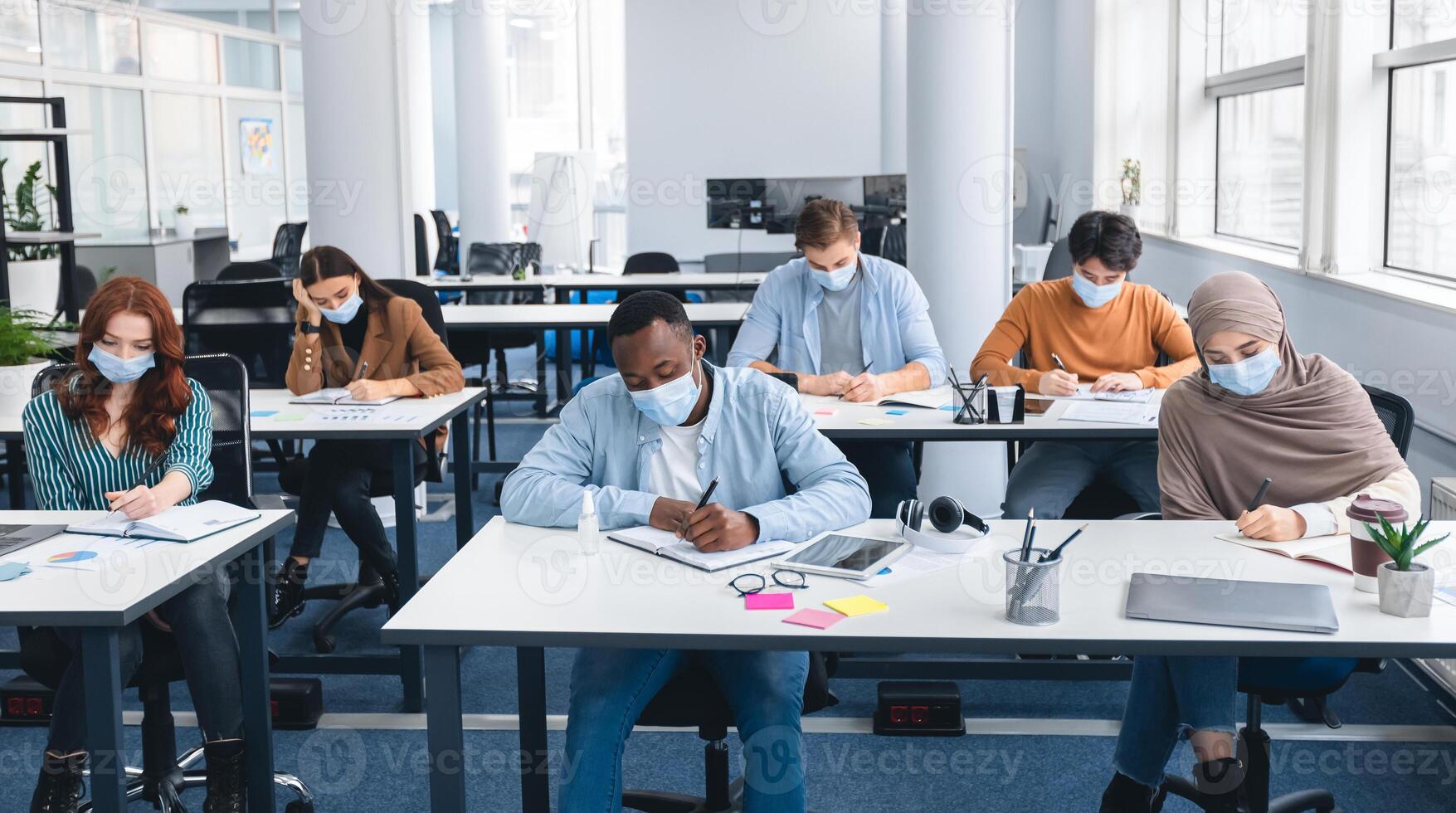 Modern Education And Studying During Pandemic Concept. Diverse group of international students sitting at desks in classroom at high school, wearing protective medical mask, writing in notebooks photo