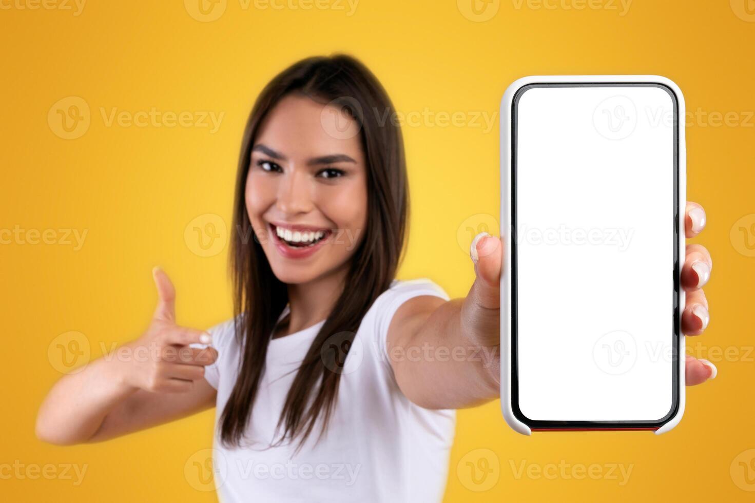 Cheerful young lady holding smartphone with white empty screen, pointing at blank display for mobile application advertisement or your text, gadget mockup template, orange studio background photo