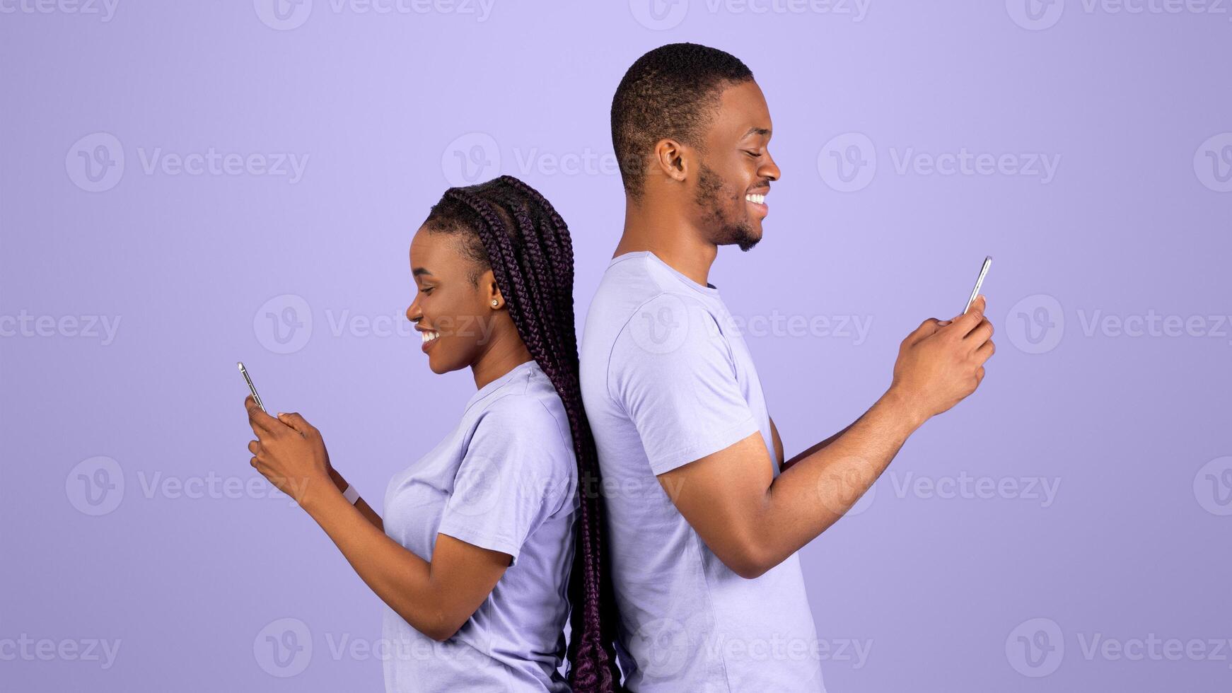 Modern Lifestyle. Profile side view portrait of cheerful positive African American couple standing back to back, holding and using their mobile phones and chatting online, violet studio background photo