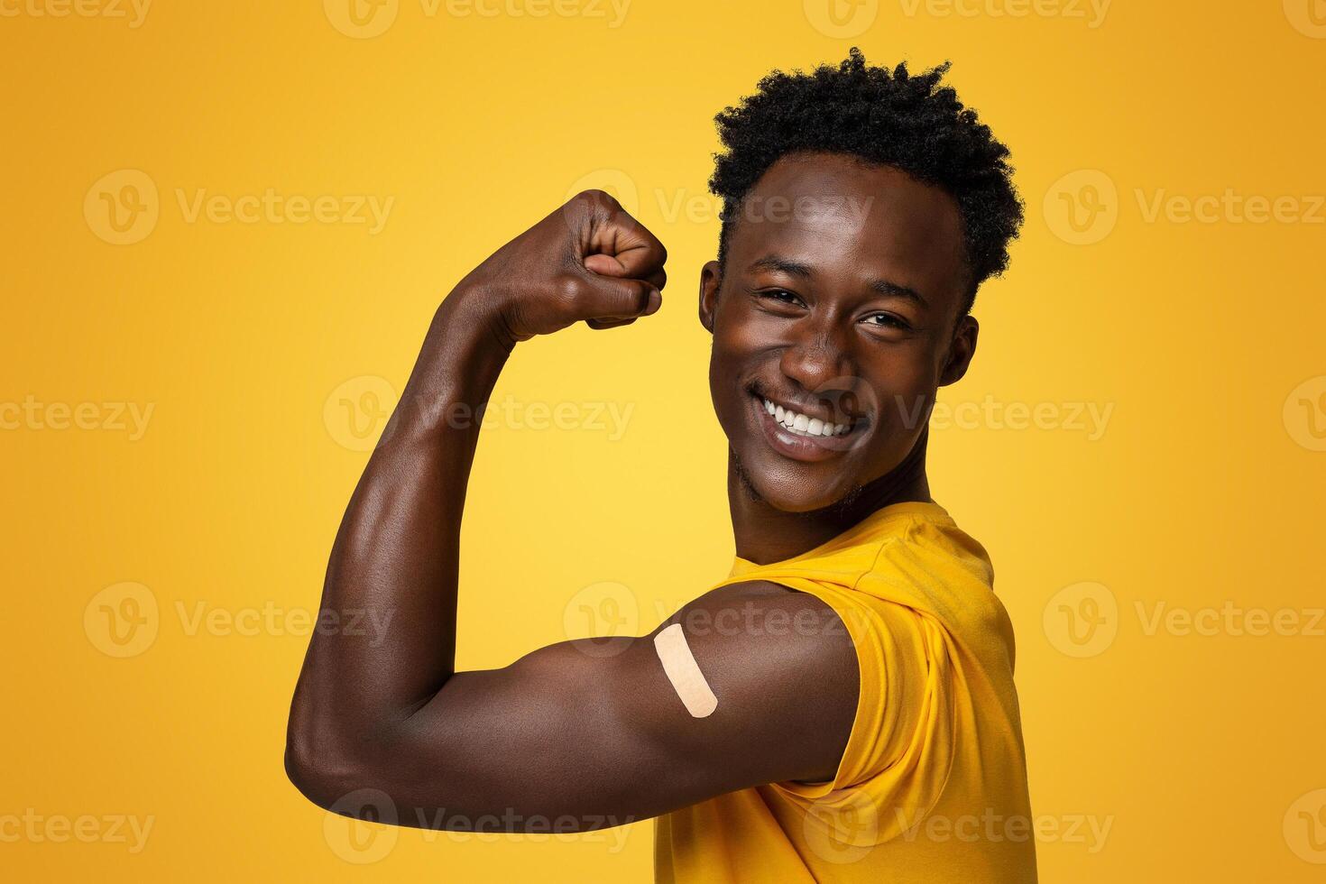 Happy Black Guy Showing Arm With Adhesive Bandage After Covid-19 Vaccination Shot, Vaccinated Young African American Man Demonstrating Biceps With Plaster, Sitting Over Beige Studio Background photo
