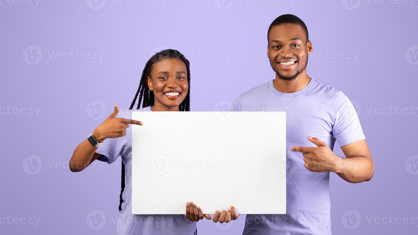 Cheerful Black Couple Holding Blank Paper Poster Together And Pointing At Free Space For Your Text Smiling To Camera Standing Over Purple Violet Studio Background. People With White Board For Advert photo