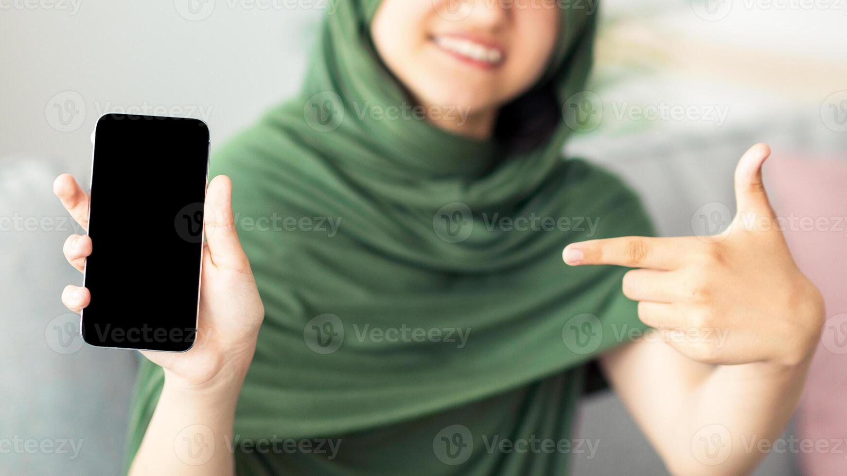 Unrecognizable Muslim teen girl in hijab pointing at cellphone with empty screen at home, mockup for design. Cheery Islamic adolescent offering smartphone with space for mobile app, closeup view photo