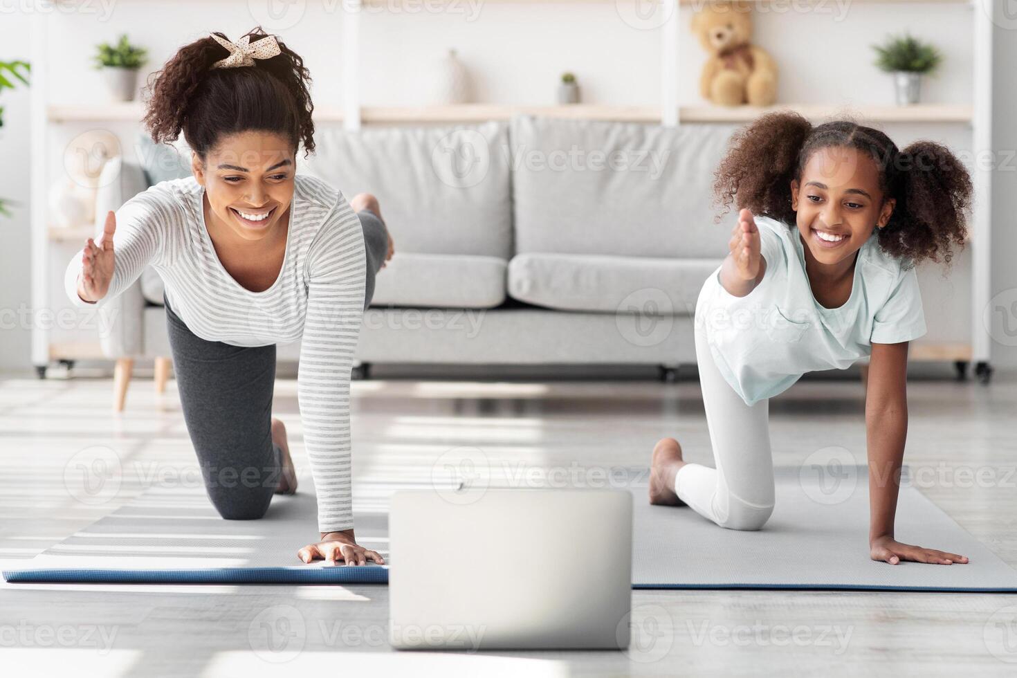 Beautiful black mom and teen daughter having online yoga class together, making exercises on fitness mat in front of laptop, living room interior. Family sport during COVID-19 quarantine photo