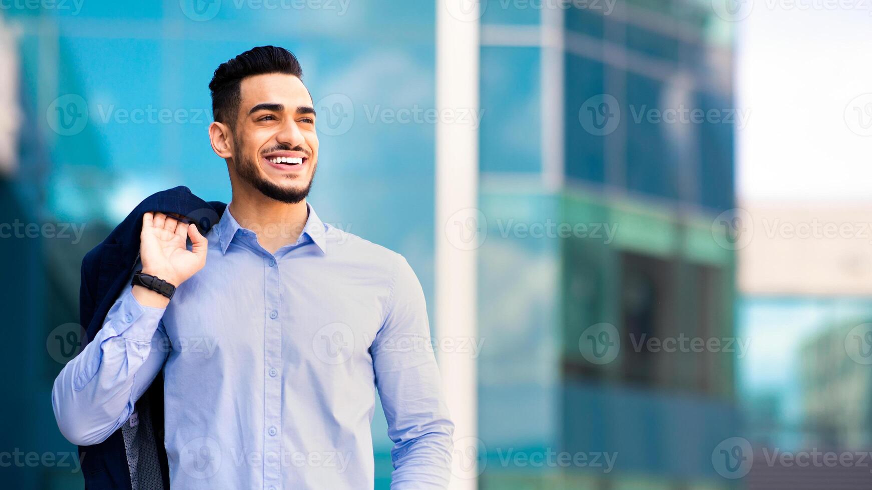 Happy young muslim man entrepreneur going home from job, carrying his jacket on shoulder, holding hand in jeans pocket, looking at copy space and smiling, posing by office building, panorama photo