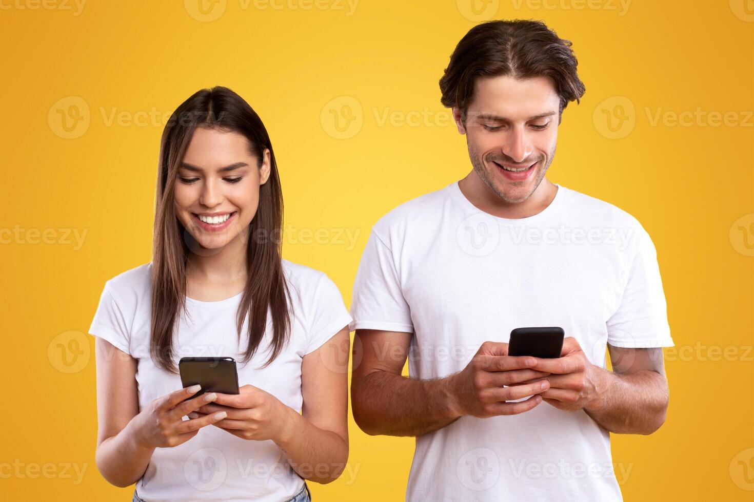 Portrait of cheerful couple holding their smartphones and playing game, texting sms or chatting online. Millennial man and woman using gadgets, standing isolated on yellow orange studio background photo