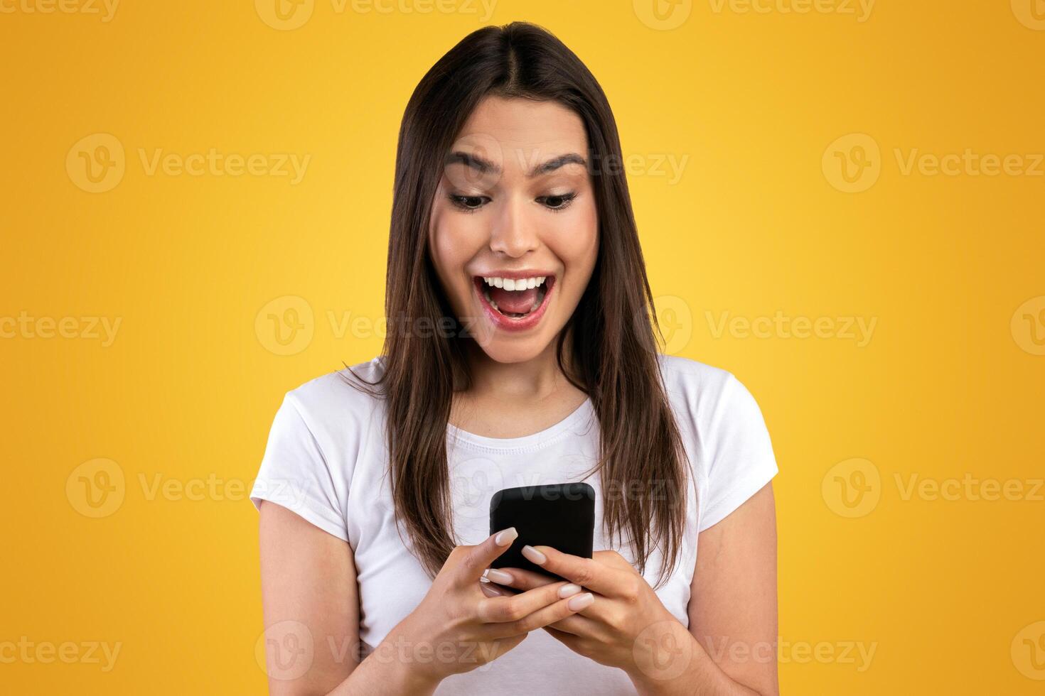 People And Technology Concept. Portrait of excited young woman using smartphone standing isolated on orange studio background, looking at cell phone screen. Lady chatting online, browsing social media photo