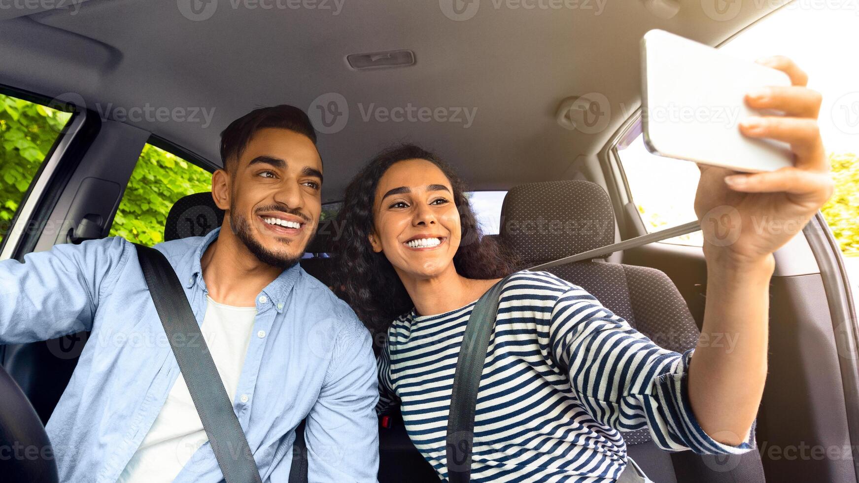 Cute cheerful middle-eastern couple driving car and taking selfie on smartphone. Happy arab millennial man and woman taking photo on mobile phone while going vacation by cozy auto