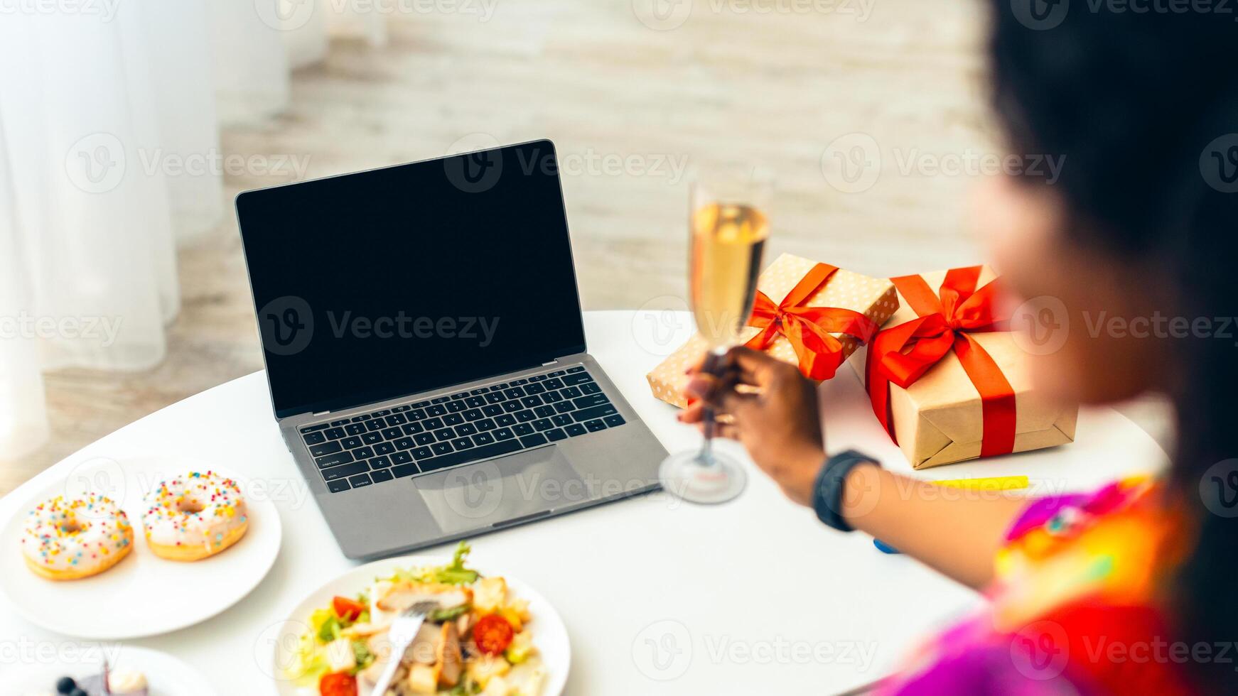 Online Holiday Celebration. Back-View Of Black Lady At Laptop With Empty Screen Celebrating Birthday Or New Year Holding Glass Of Wine, Sitting At Table With Salad, Donuts And Gifts Boxes Indoor photo