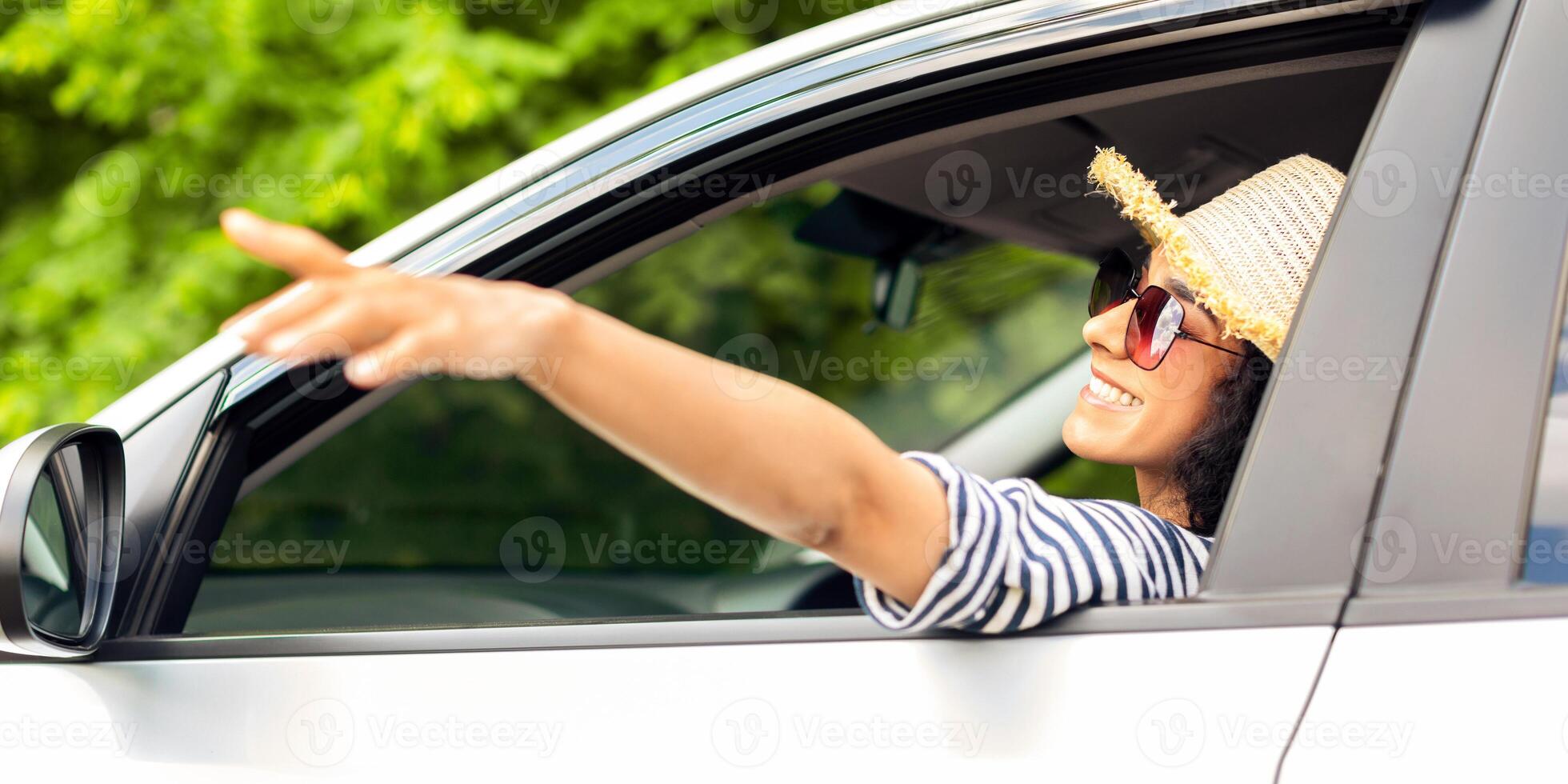 Happy middle-eastern young woman in stylish summer hat and sunglasses enjoying car trip, sitting at front seat in nice auto, looking through window, touching air with hand, side view photo