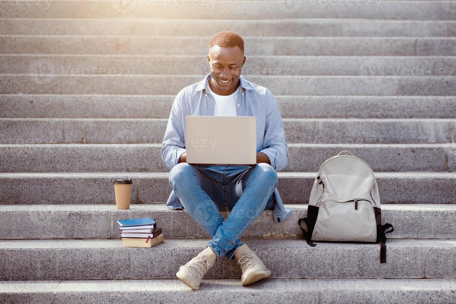 Focused black guy studying online with laptop computer on stairs downtown photo